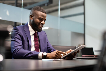 Cheerful millennial black guy in nice suit CEO sitting at table, using digital tablet at office, checking newest business mobile application and smiling, copy space. Modern technologies and business