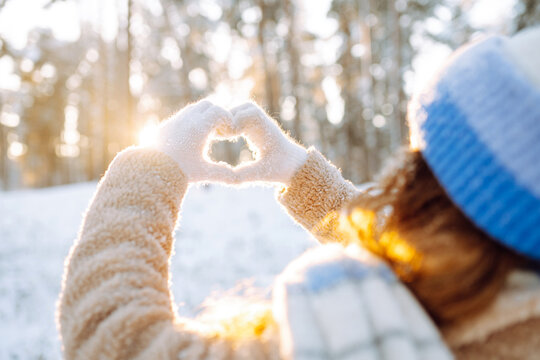Rear view of woman in cozy winter clothing stands in snowy forest, clasping hands in heart shape. Beautiful woman enjoys sunny winter day in park. Conceptual concepts of symbolism, seasonality.