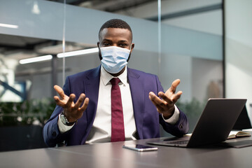 Young black manager in expensive blue suit and face mask working at office while COVID-19 pandemic, sitting at table with laptop, gesturing, having conference with business partners, copy space