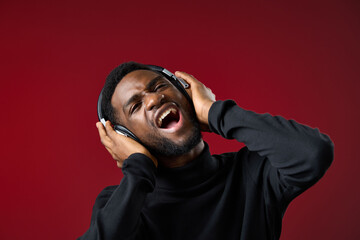 Young African man enjoying music while singing passionately with headphones on, vibrant red background, showcasing emotion and energy