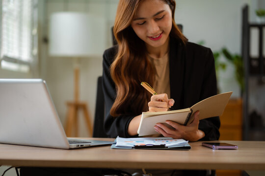 Female hand taking notes on a notebook handwritten note writing business schedule and use a laptop computer at the office desk in the morning.