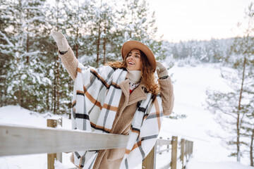 Cute woman wrapped in a warm scarf and wearing a stylish hat, stands on a wooden path on a snowy...