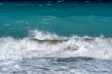 Powerful turquoise sea wave crashing on pebble shore with white foam in sunlight. Dynamic ocean surf background for summer, travel and nature concepts.