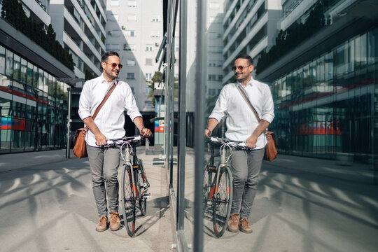 Young man walking bicycle beside modern building reflecting image