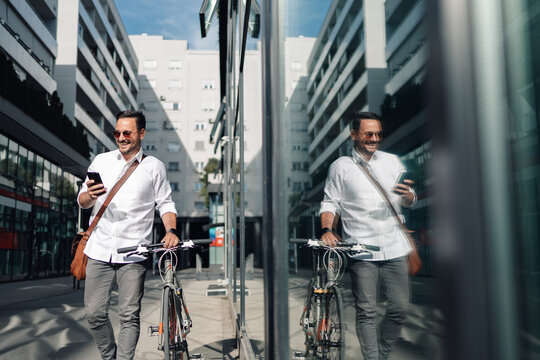 Businessman walking with bicycle checking smartphone in urban setting - Powered by Adobe
