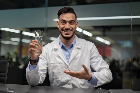 A young middle eastern doctor in workwear smiles at the camera while holding a glass of water. He promotes the importance of regular water intake for a healthy lifestyle in a bright office setting.