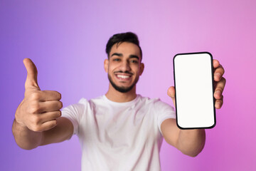 Young Arab man stands in neon-lit setting, smiling widely while holding a modern smartphone with a blank white screen. He gives a thumbs up, promoting an app or website design.