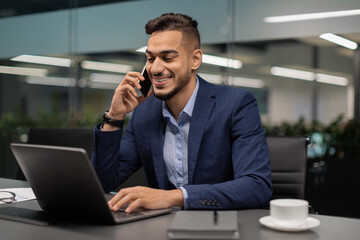 Middle eastern businessman sits at a modern office table, happily talking on the phone with a client. He types on his laptop and enjoys a cup of coffee during a productive workday.
