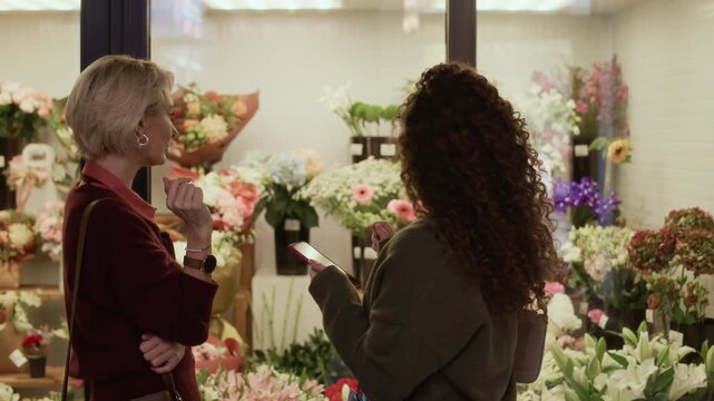 Medium shot of two Caucasian female friends looking at display with flower bouquets and taking photos in florist shop