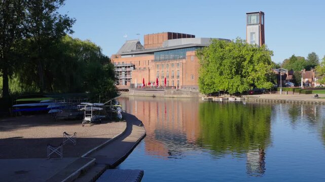 A static wide shot of the modern Royal Shakespeare Theatre and its iconic tower reflecting clearly in the River Avon in Stratford-upon-Avon on a sunny autumn morning.