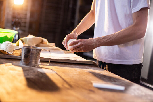 Hands of person shaping dough on wooden surface, preparing food in bakery setting, warm light casting shadows on counter, culinary craftsmanship - Powered by Adobe