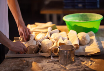 Baker hands preparing bread dough for baking process. Raw bread forms resting on floured linen cloths on wooden table. Rustic bakery scene with fresh dough and essential baking tools.