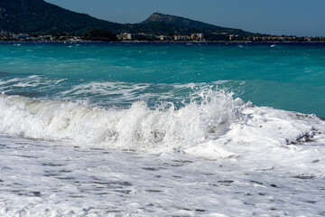 Turquoise Mediterranean sea wave crashing on sandy beach with white foam, coastal town and mountain ridge in background. Summer seaside travel landscape.