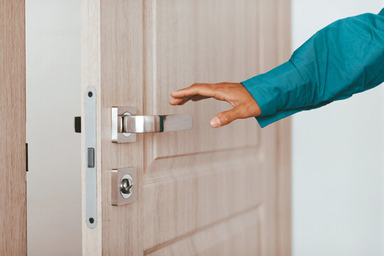 A hand positioned to turn the silver handle of a wooden door, showcasing a clean interior with natural light. The moment suggests intentionality, as the door leads to another space.