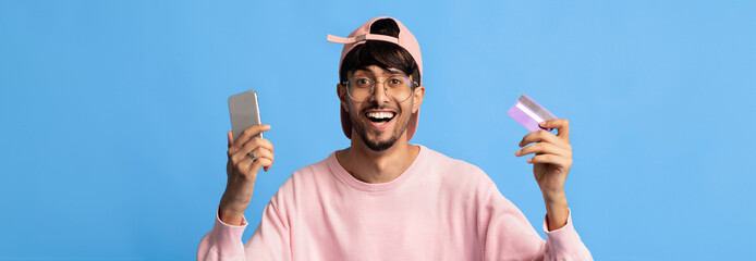 A cheerful young man holds a smartphone in one hand and a card in the other. He wears a pink sweater and cap, posing against a vibrant blue background that adds to his lively expression.