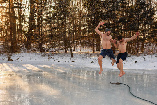 Two happy barefoot men in swimwear running through a sprinkler on a frozen lake holding hands