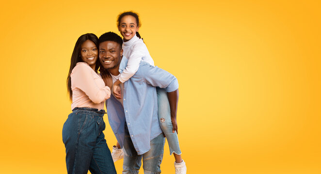 A joyful young african american family poses together with a smiling father giving his daughter a piggyback ride. They are standing against a bright yellow background, radiating happiness.
