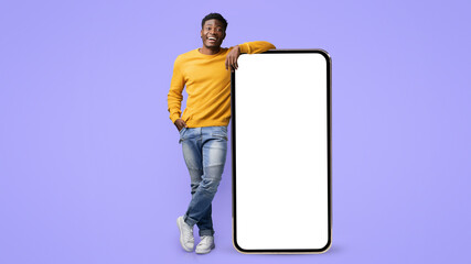 A man wearing a yellow sweater and blue jeans stands confidently, leaning on a large blank mobile device. The vibrant purple background adds a fun touch to the scene.