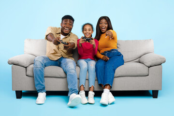 A joyful African American family is sitting on a couch, playing video games and using controllers. They are smiling and having fun in a blue studio background during their free time.