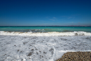 Turquoise sea wave with white foam washing over pebble beach under clear blue sky and distant islands on horizon