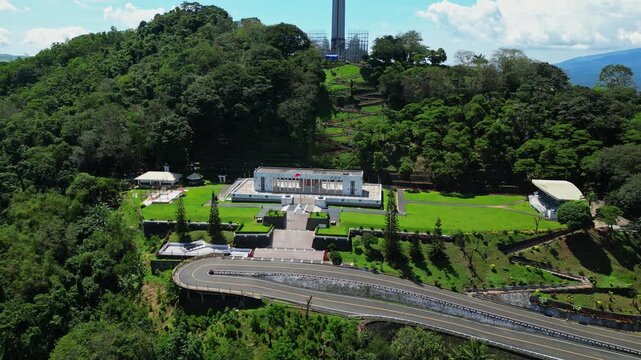 Tilt‑down aerial revealing the museum at the base of Mt. Samat National Shrine, framed by manicured lawns, pathways, and lush forested hills in Pilar, Bataan, Philippines.