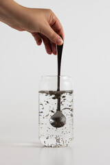 Stirring chia seeds from a silver spoon into a glass of water on a white background