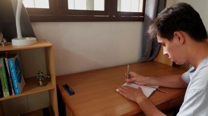Static medium shot of a young man seated at a wooden desk, writing in an A5 notebook journal by a sunlit window.