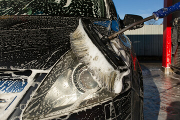 Self service car wash scene showing a soapy brush scrubbing the front headlight of a black vehicle, with foam, bubbles and reflections visible on the wet body surface.