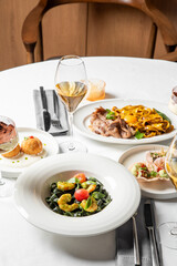 A high-angle vertical shot of a white table at a fine dining restaurant, set with various delicious Italian pasta dishes and glasses of white wine