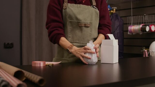 Cropped shot of unrecognizable gift shop worker in apron wrapping vase in paper and putting in box at working table