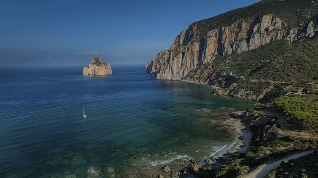 Aerial view of the iconic Pan di Zucchero rock formation rising from the turquoise sea, framed by rugged cliffs and lush greenery, Masua, Sardinia, Italy.
