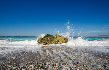 Turquoise sea waves crashing against lonely rock on pebble beach, white foam splashing high under clear blue summer sky.