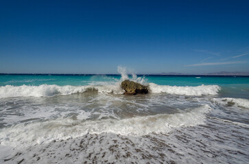 Turquoise sea waves crashing against lonely rock on pebble beach, white foam splashing high under clear blue summer sky.