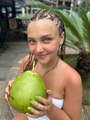 Young woman holding a fresh green coconut in front of lush tropical greenery. Vibrant summer atmosphere, travel lifestyle, exotic vacation mood, nature background.