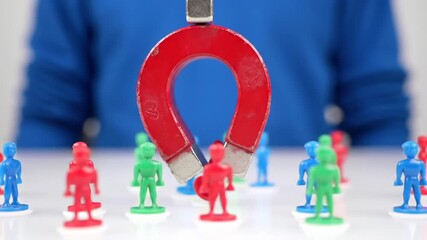 A red horseshoe magnet attracts a red figure from a group of plastic figurines in blue, red, and green. The background is a blurred figure in blue on white.