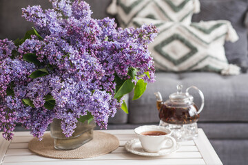 The concept of home comfort. A large purple bouquet of lilacs, a cup of tea, and a teapot on the table. In the background is the interior of a modern living room.