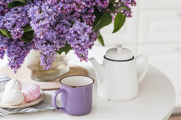 The concept of home comfort. A large purple bouquet of lilacs, a ceramic teapot, a cup and a delicious marshmallow on the table. In the background is the interior of a modern white kitchen.