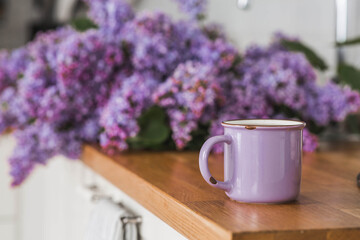 The concept of home comfort and drinks. A lilac cup of tea or coffee on the kitchen countertop. In the background is a large purple bouquet of lilacs.