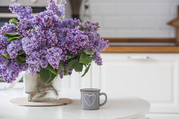 The concept of home comfort. A large purple bouquet of lilacs and a cup of tea on the table. In the background is the interior of a modern white kitchen.