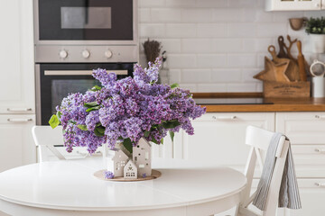 The concept of home comfort. A large purple bouquet of lilacs and white Scandinavian houses on the table. In the background is the interior of a modern white kitchen.