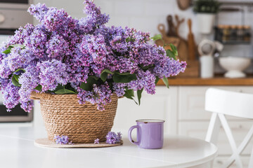 The concept of home comfort and celebration. A large purple bouquet of lilacs in a wicker basket, a teapot and a cup of tea on the table. In the background is the interior of a modern white kitchen.