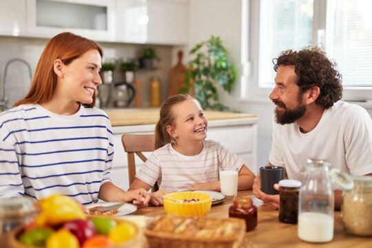 A cheerful family sits at a wooden table enjoying breakfast together. The mother, father, and daughter share laughs while surrounded by fresh fruits and beverages, creating a cozy environment.