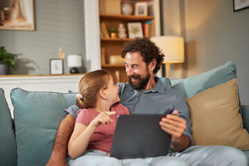 A father and his young daughter share a joyful moment on a cozy couch, interacting with a tablet and smiling at each other in their family living room.