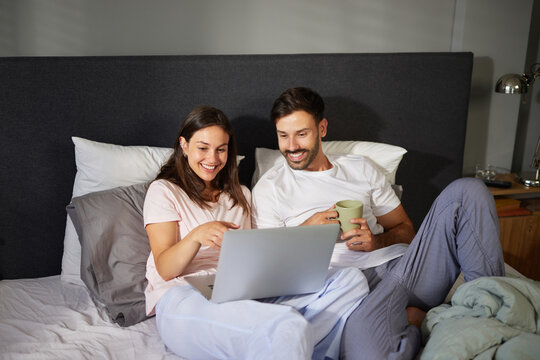 Two people are having a relaxing evening together in bed, smiling and engaging with a laptop while sipping warm beverages in a cozy environment.