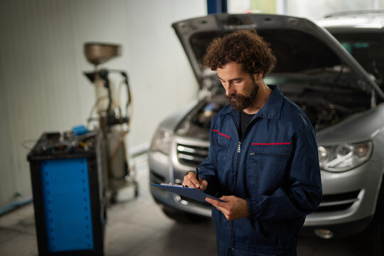 A mechanic in a blue uniform reviews vehicle details on a clipboard in a busy repair shop. The car hood is open, revealing the engine compartment. Tools are visible nearby. - Powered by Adobe