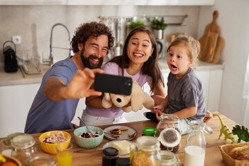 Happy family captures a joyful moment during breakfast at home. Parents and child pose with smiles, with a soft toy in hand, creating lasting memories together.