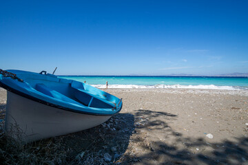 Blue fishing boat chained on sandy beach with turquoise sea waves and clear blue summer sky, Mediterranean coastal landscape, travel and vacation concept.