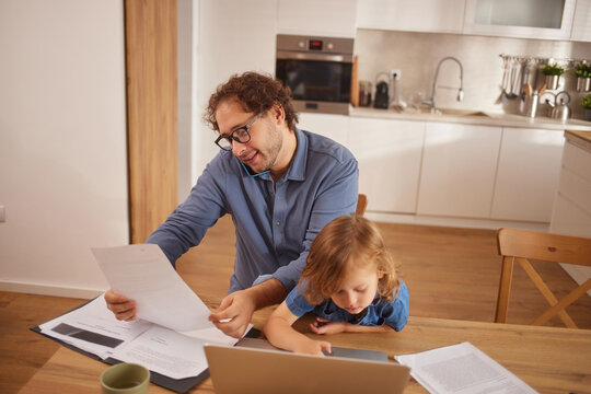 A man reviews paperwork while talking on the phone, assisting a young child with a laptop. Their kitchen is bright, reflecting an enjoyable family time.