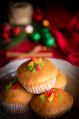 A warm and festive still life of Christmas-themed cupcakes topped with colorful candied fruit. The cupcakes are arranged with holiday decorations including pine branches, red berries, and rustic.