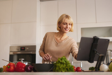 In a bright kitchen, a woman smiles as she cooks, surrounded by fresh vegetables and a tablet displaying a recipe. She enjoys making a nutritious meal.